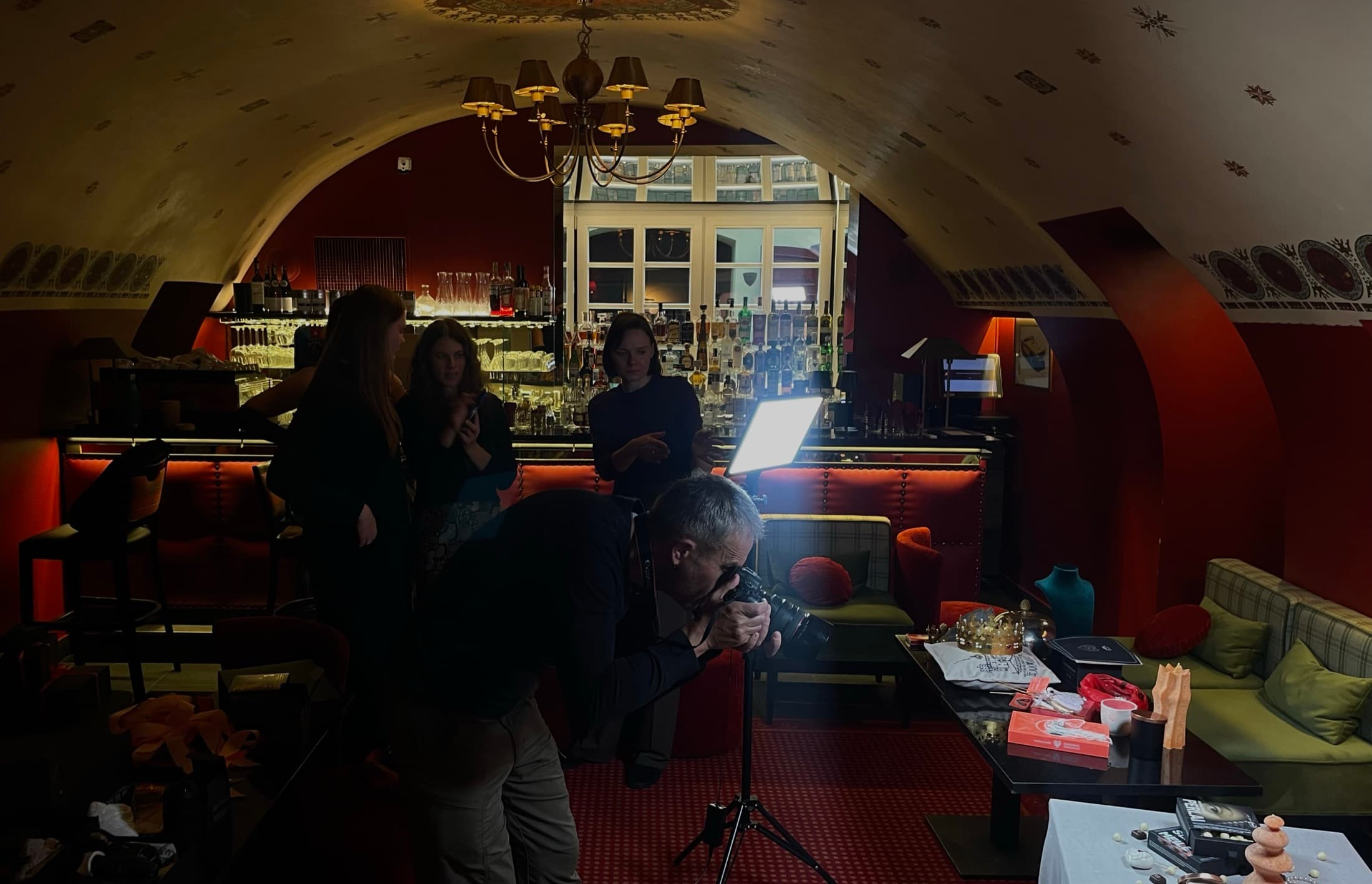 A photographer crouched over a camera on a tripod in a red vaulted bar, a lighting panel and crew behind him