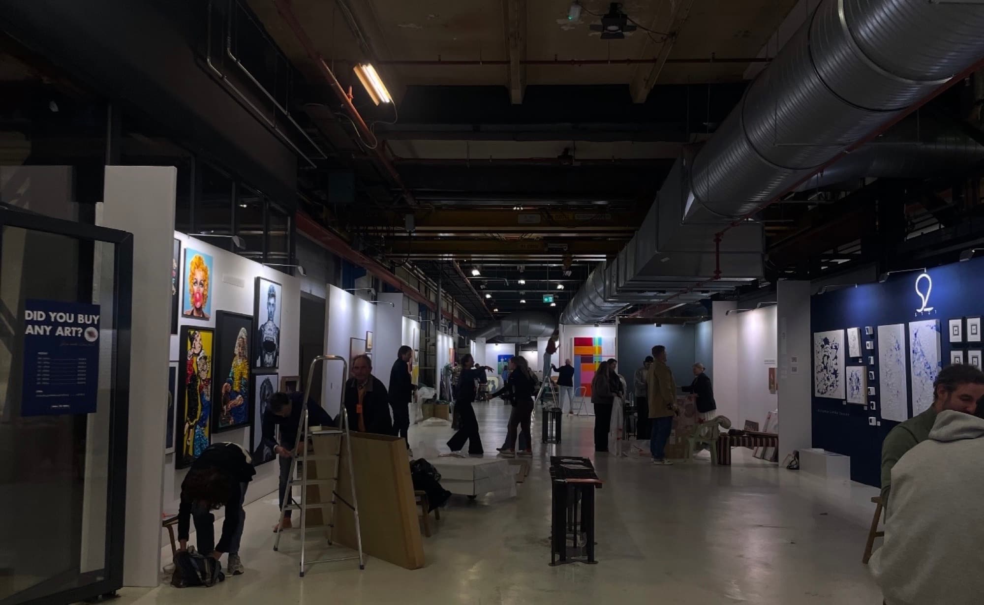 An art fair hall during build-up, stands going up under exposed ducting